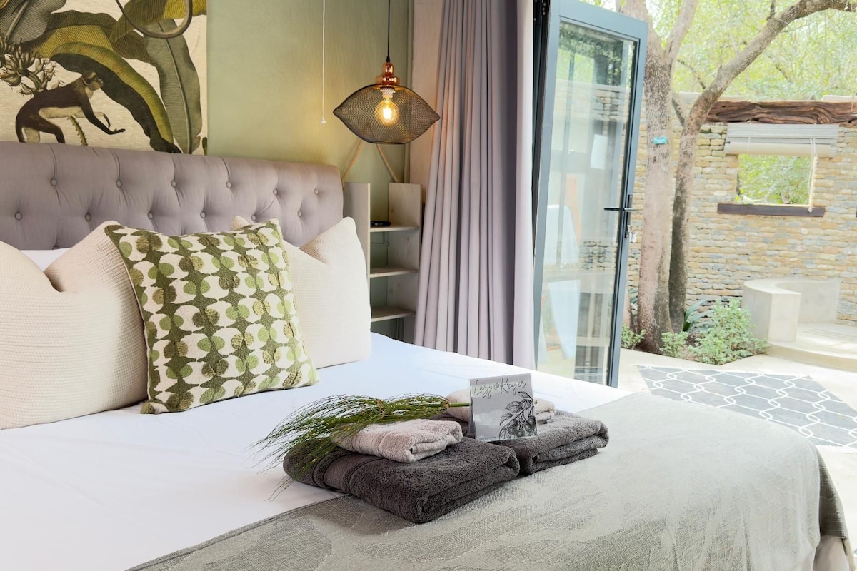 Un dormitorio con cabecera capitonada, cama con almohada estampada, lámpara de noche y vistas a un espacio exterior con muro de piedra y vegetación.