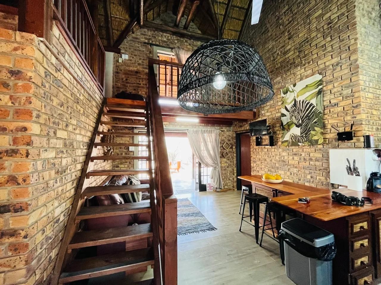 A rustic interior with exposed brick walls, a wooden staircase, a dining area with bar stools, and a hanging light fixture.
