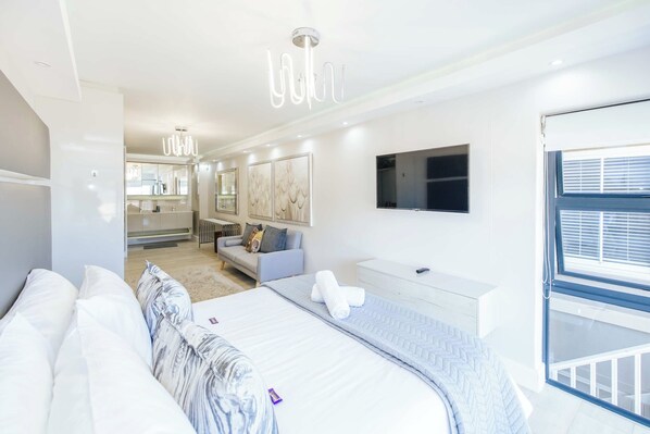 Une chambre moderne avec un grand lit, un canapé, une télévision et une salle de bain visible au fond.