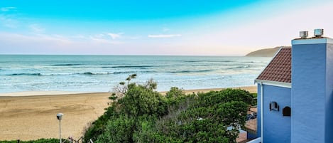 A coastal view with a beach, a clear sky, and a building with a red-tiled roof.