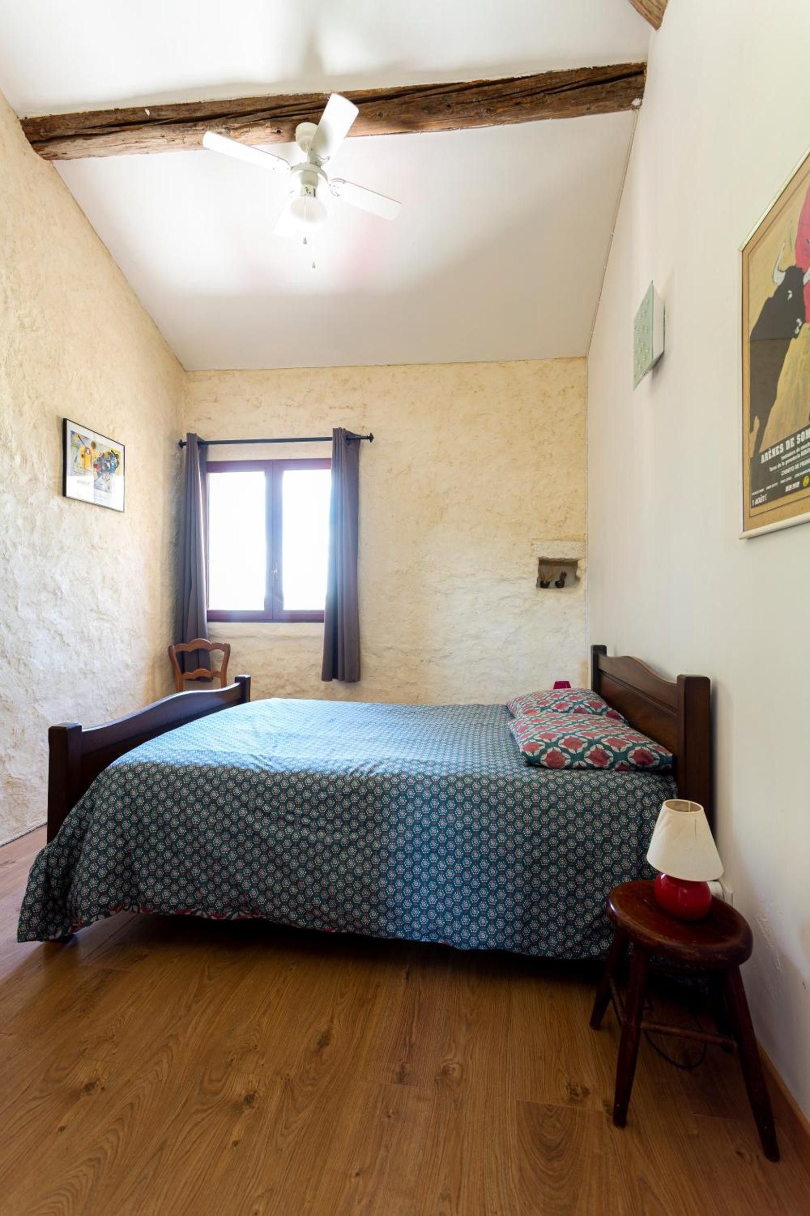 Een slaapkamer met een bed, houten vloer, een plafondventilator, een raam met gordijnen, een stoel en een klein tafeltje met een lamp.