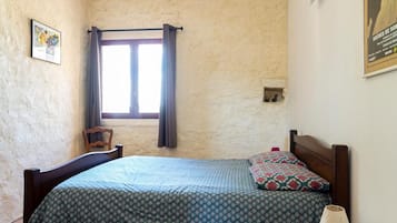 Een slaapkamer met een bed, houten vloer, een plafondventilator, een raam met gordijnen, een stoel en een klein tafeltje met een lamp.