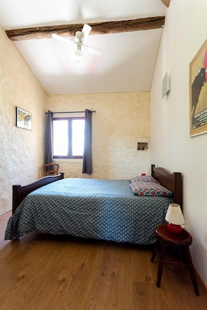 A bedroom with a bed, wooden floor, a ceiling fan, a window with curtains, a chair, and a small table with a lamp.