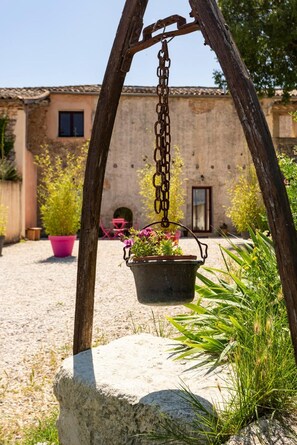 A rustic outdoor area with a hanging metal pot containing flowers, a stone bench, and a building with a tiled roof in the background. - Cottage 'Mas Du Cres Gîte' with Private Terrace, Private Garden and Wi-Fi (Galargues)