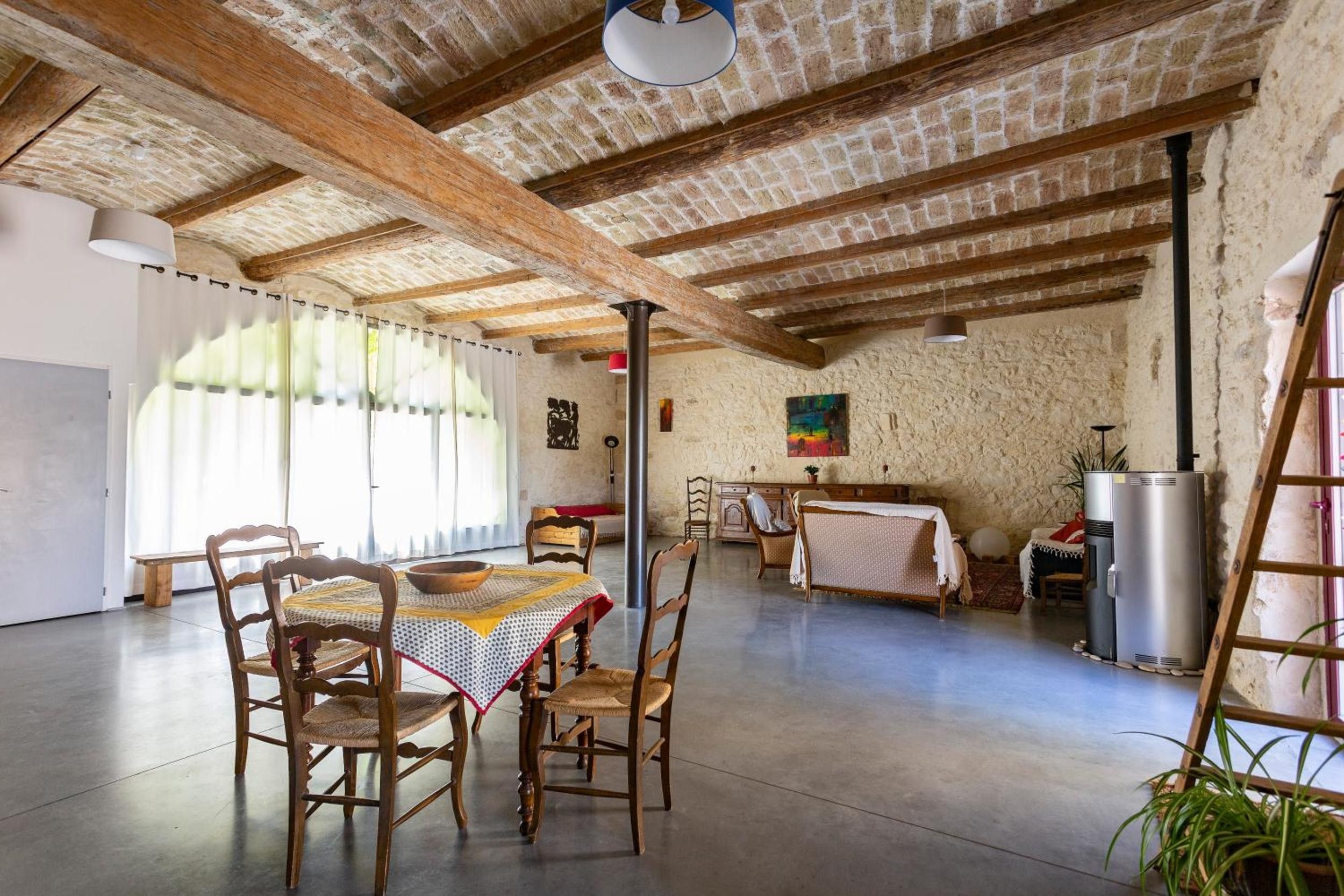 Een ruime kamer met een hoge nok, zichtbare houten balken, een eettafel met stoelen, een bed, een radiator en een raam met gordijnen.