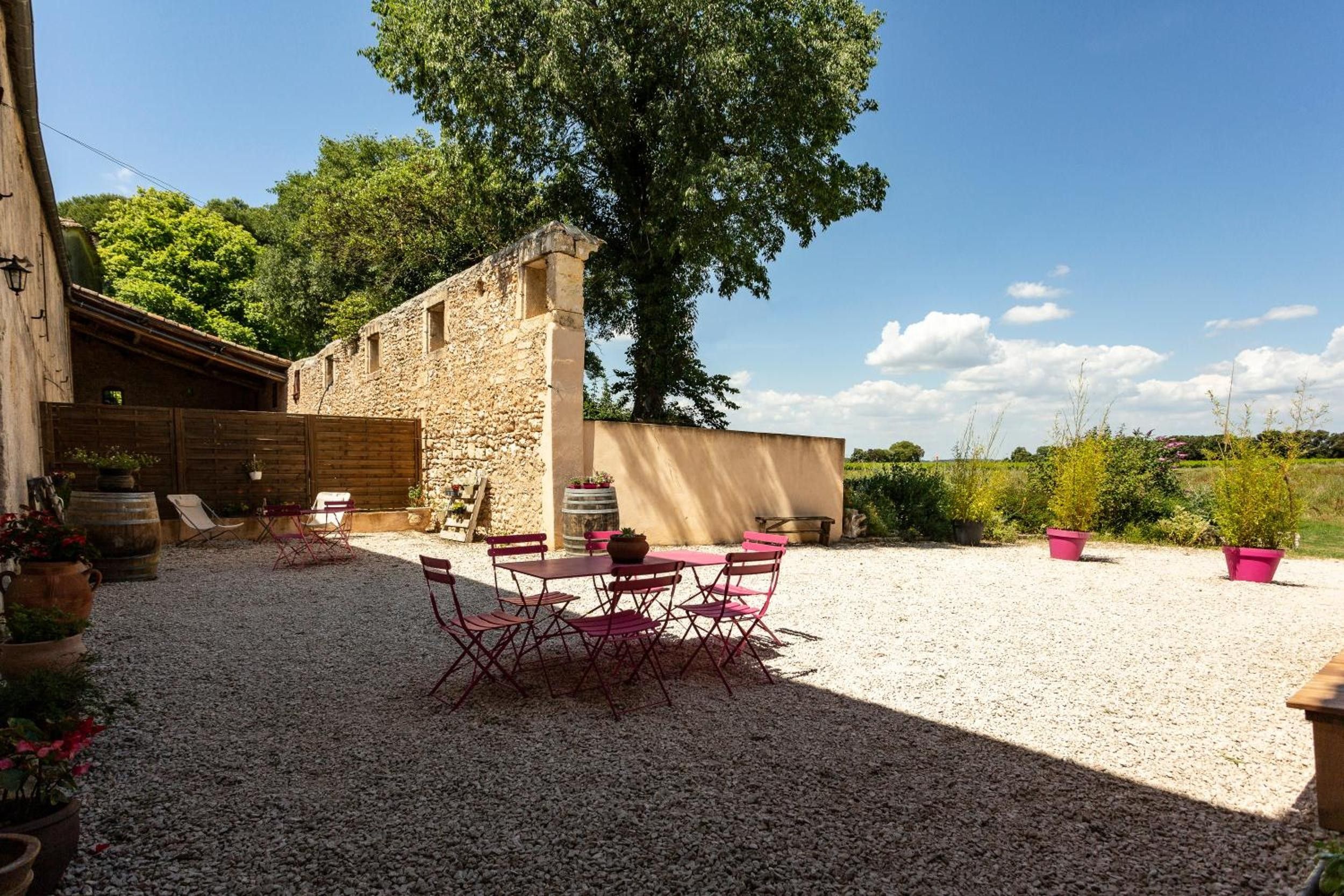 A rustic outdoor seating area with a stone building, a large tree, and gravel ground.