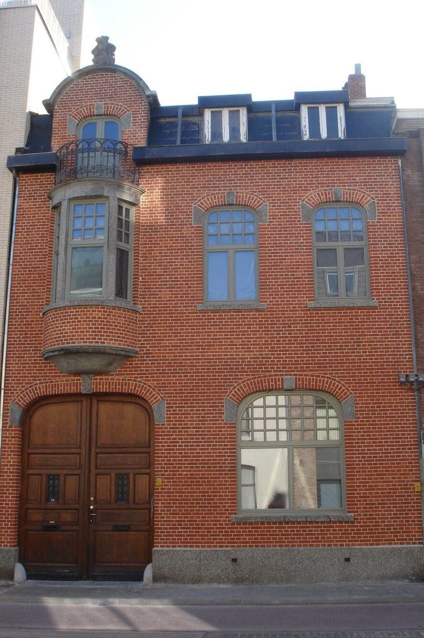 A red brick building with a large wooden double door, two windows with arched tops, and a small balcony with a metal railing.