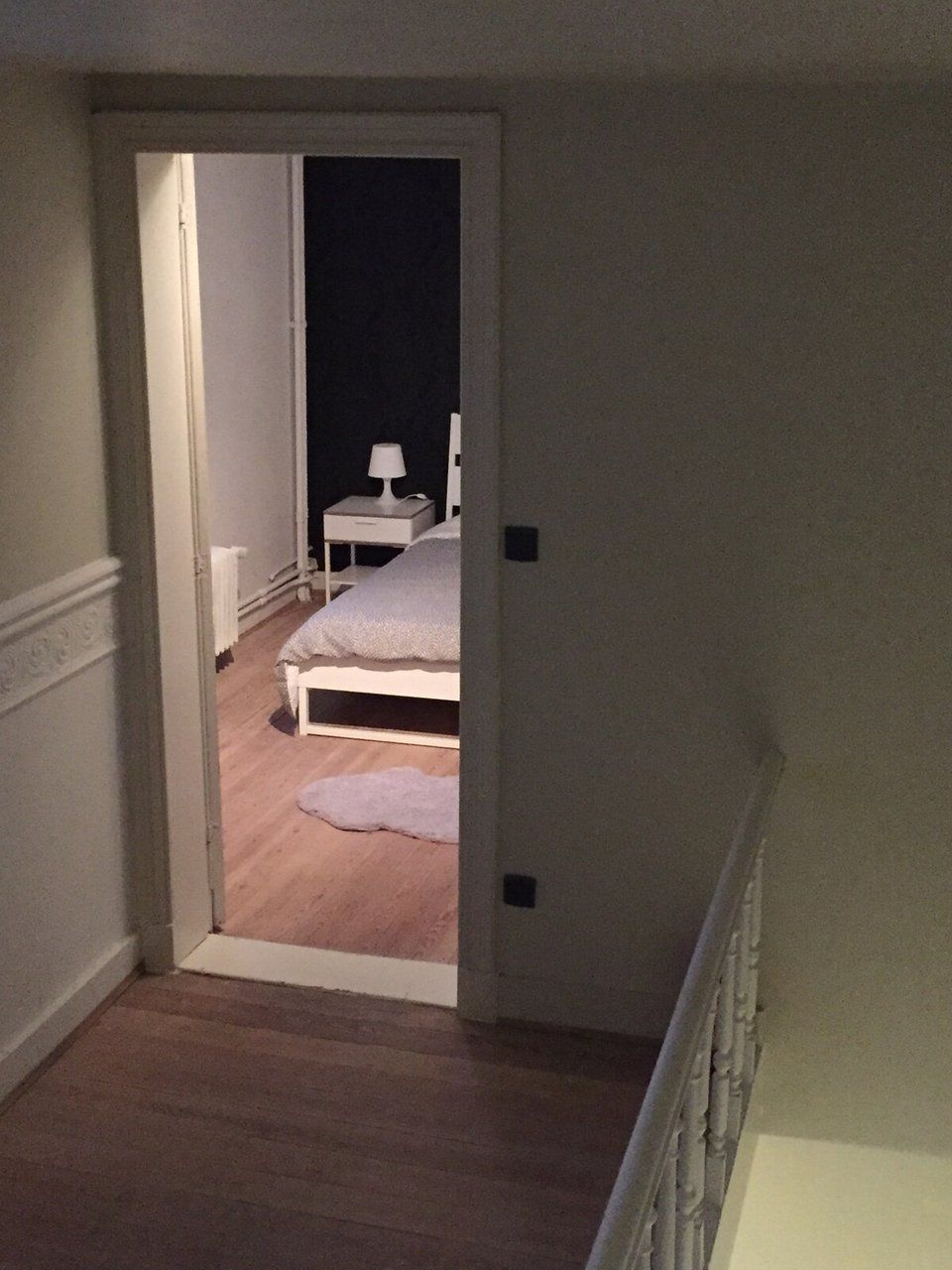 Een smalle gang die leidt naar een slaapkamer met een bed en nachtkastje.