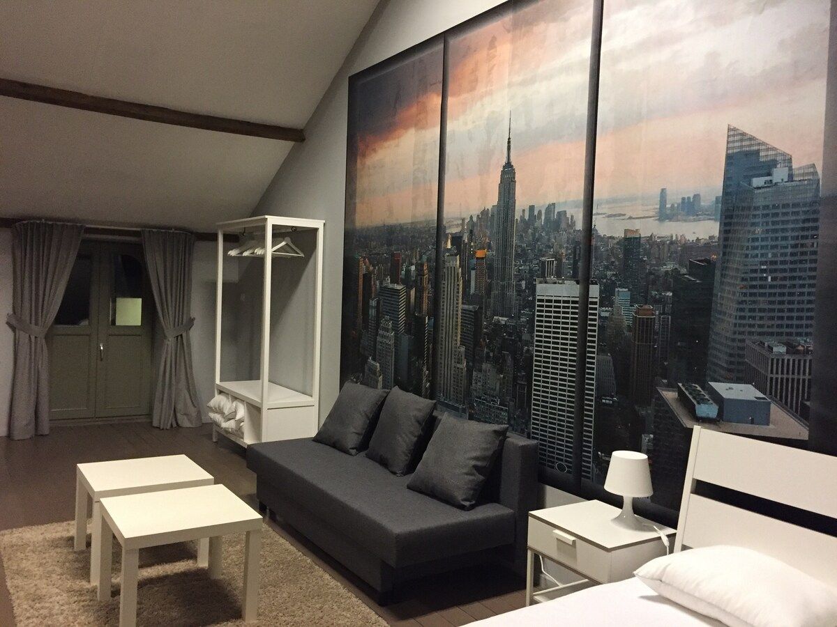 A hotel room with a large cityscape mural, a sofa, a bedside table, a lamp, and a wardrobe.