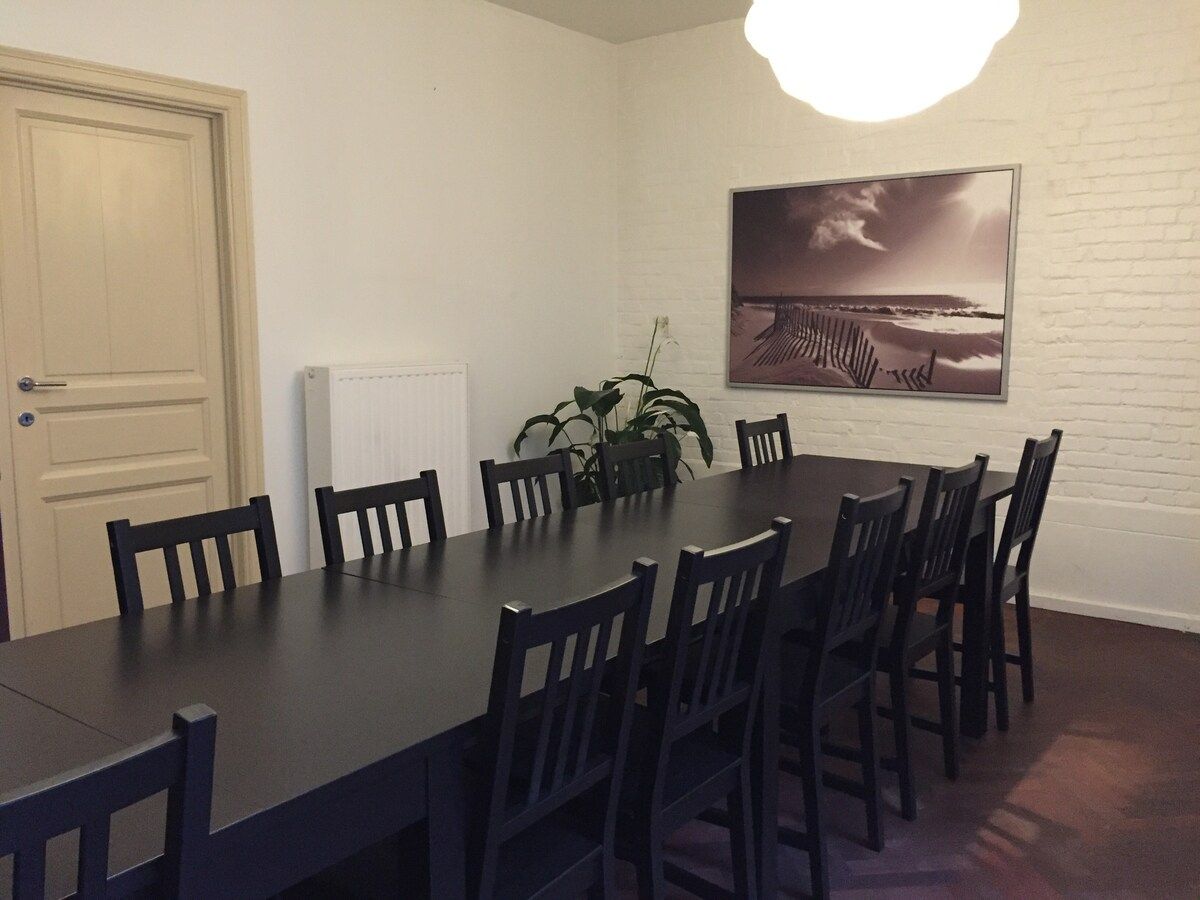 Een eetgedeelte met een lange tafel en stoelen, een plant en een schilderij aan de muur.