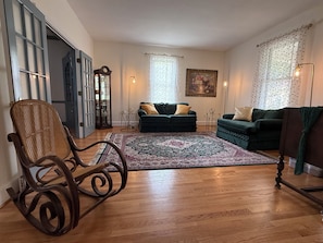 Un salon avec un plancher en bois, un fauteuil à bascule, deux canapés verts, un tapis à motifs et un tableau accroché au mur.