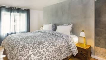 A bedroom with a bed, two bedside lamps, a large grey wall, and a window with curtains.