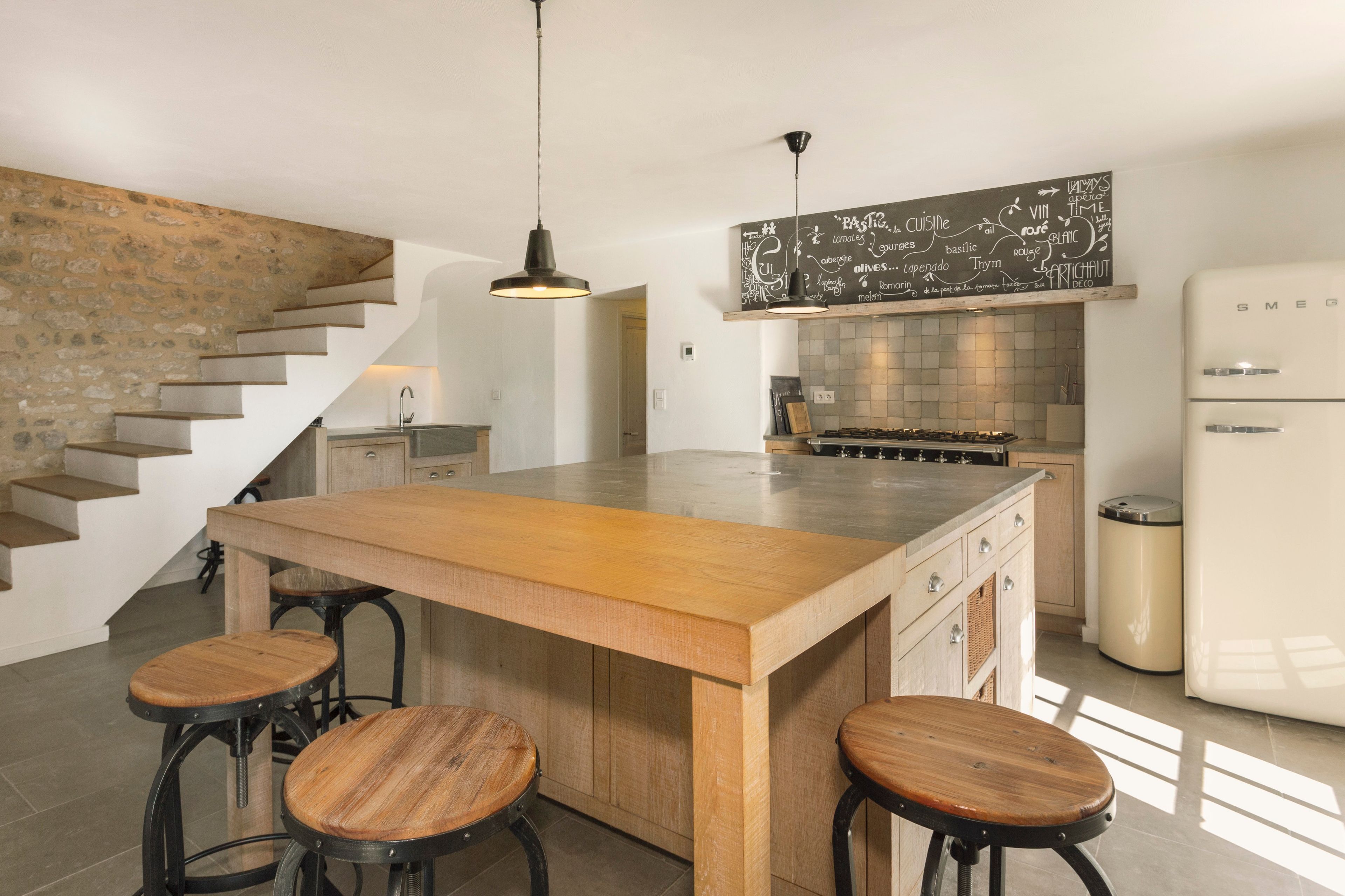 Une cuisine moderne dotée d’un îlot central, de tabourets de bar, d’un luminaire suspendu et d’un tableau noir sur lequel sont écrits des termes culinaires.