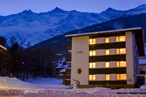 Ein Hotel mit einem Schild mit dem Schriftzug 'Alfa' an der Fassade, umgeben von schneebedeckten Bergen und Bäumen.