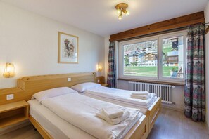 A hotel room with two beds, a window with a view of buildings, a radiator, and a wall-mounted light fixture. - Comfortable 3 Bedroom Apartment with Mountain Views Haus Alfa (Saas Fee)