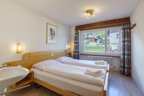 A hotel room with a wooden bed, a bedside table with a sink, a framed picture on the wall, and a window with curtains. - Comfortable 3 Bedroom Apartment with Mountain Views Haus Alfa (Saas Fee)