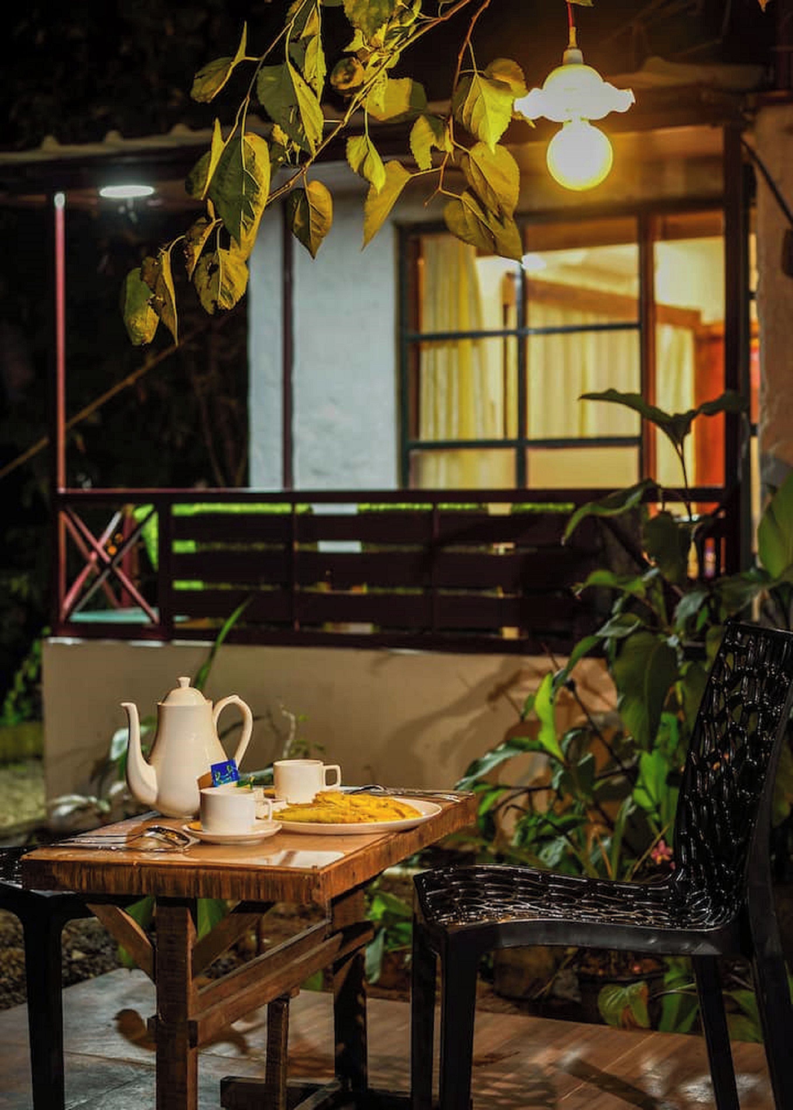An outdoor dining area with a table set for two, a teapot, cups, and a plate of food. There is a black chair and a wooden bench in the background.