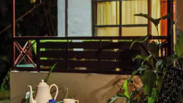 An outdoor dining area with a table set for two, a teapot, cups, and a plate of food. There is a black chair and a wooden bench in the background.