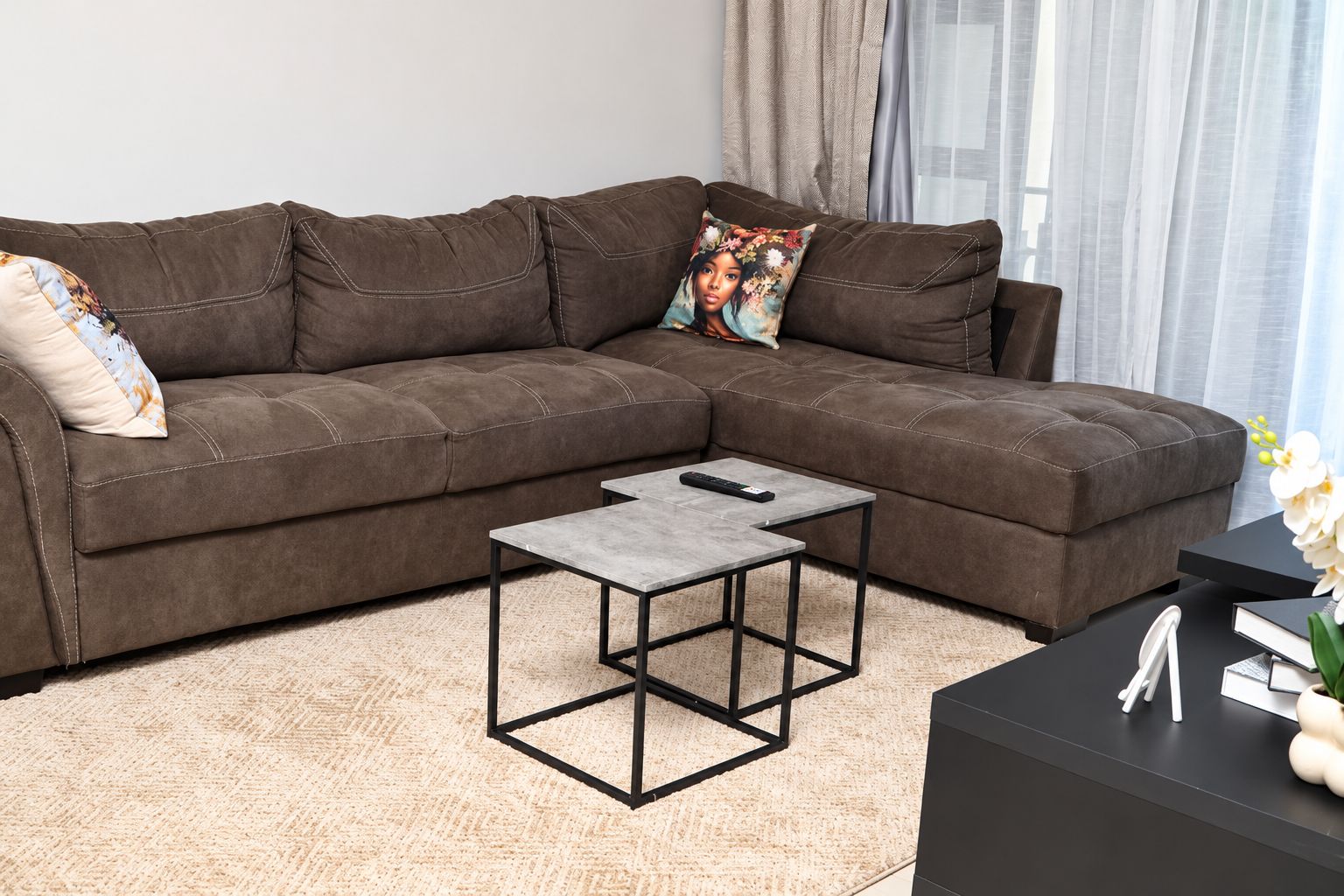 A brown sectional sofa with a decorative pillow, a small coffee table with a remote control, and a black side table with a book and a plant.