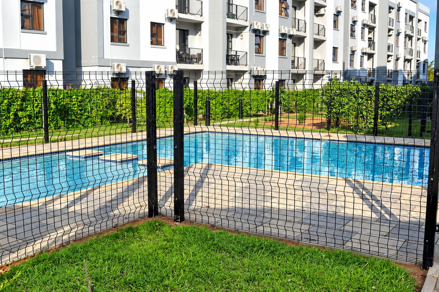 Una piscina con una valla de alambre, rodeada de césped y un edificio al fondo.