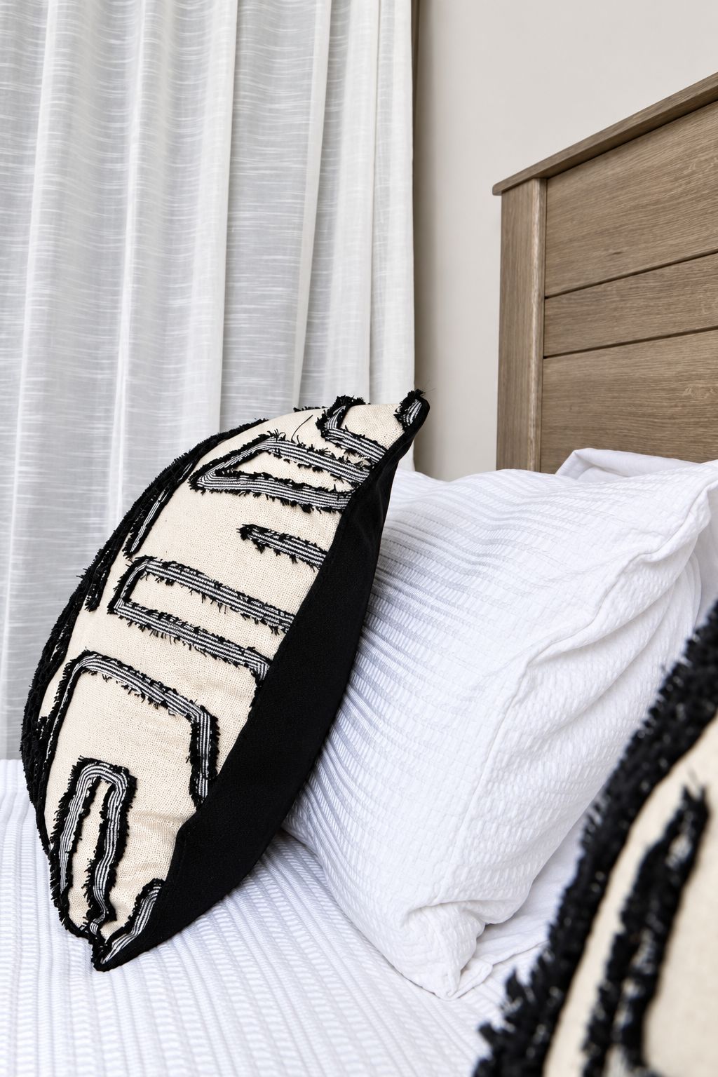 Una cama con ropa blanca y un cojín negro y beige con estampado geométrico.