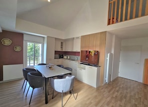 Eine moderne Küche mit weißen Schränken, ein Essbereich mit Tisch und Stühlen und ein Blick ins Freie durch ein Fenster.