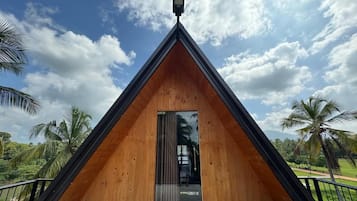 Una estructura de madera triangular con una puerta y una ventana pequeña, rodeada de palmeras y un cielo despejado.