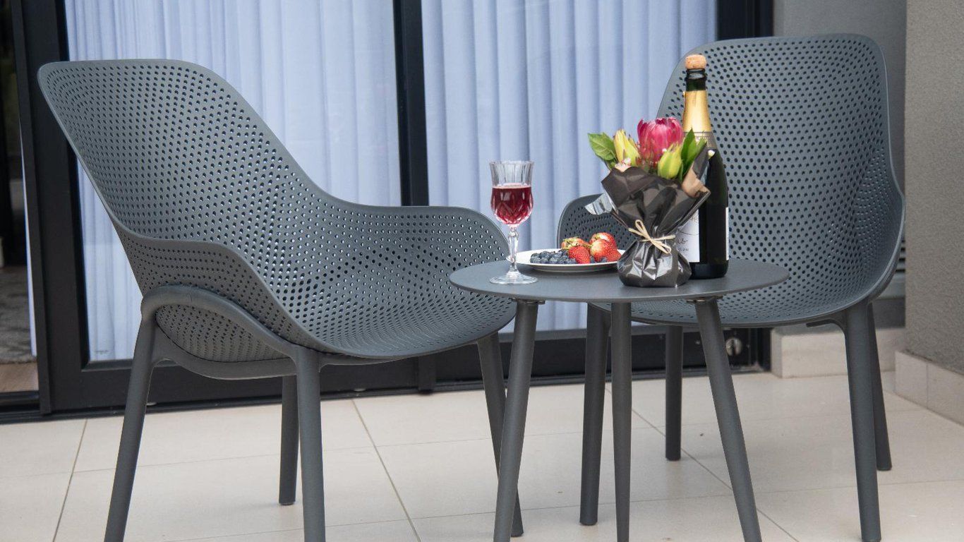 Twee metalen stoelen met gaatjes en een klein, rond tafeltje met champagne, een glas rode wijn, een bloemenboeket en een bord aardbeien.