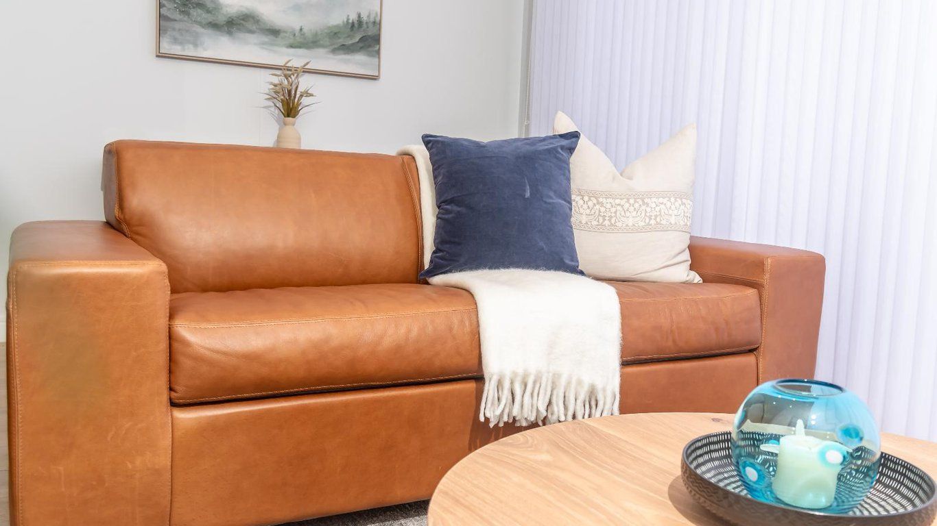 Een bruine leren bank met een witte plaid en decoratieve kussens, een ronde houten salontafel met een glazen vaas, en een lijst met een foto aan de muur.