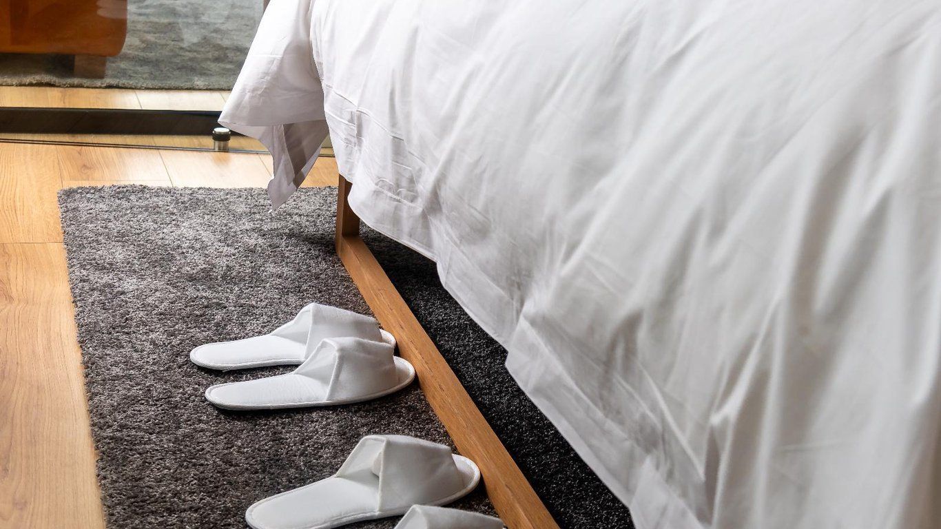 Un lit avec une literie blanche, un tapis gris foncé et des chaussons blancs posés sur le tapis.