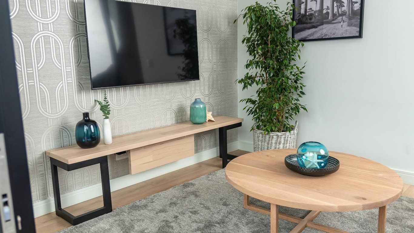 Een moderne woonkamer met een flatscreen televisie aan de muur, een houten bankje, een ronde houten tafel, een potplant en een decoratieve glazen schaal.