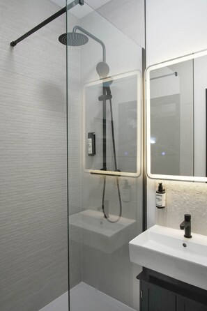 Casa de banho moderna com chuveiro em cabine de vidro, lavatório branco e espelho com iluminação embutida.