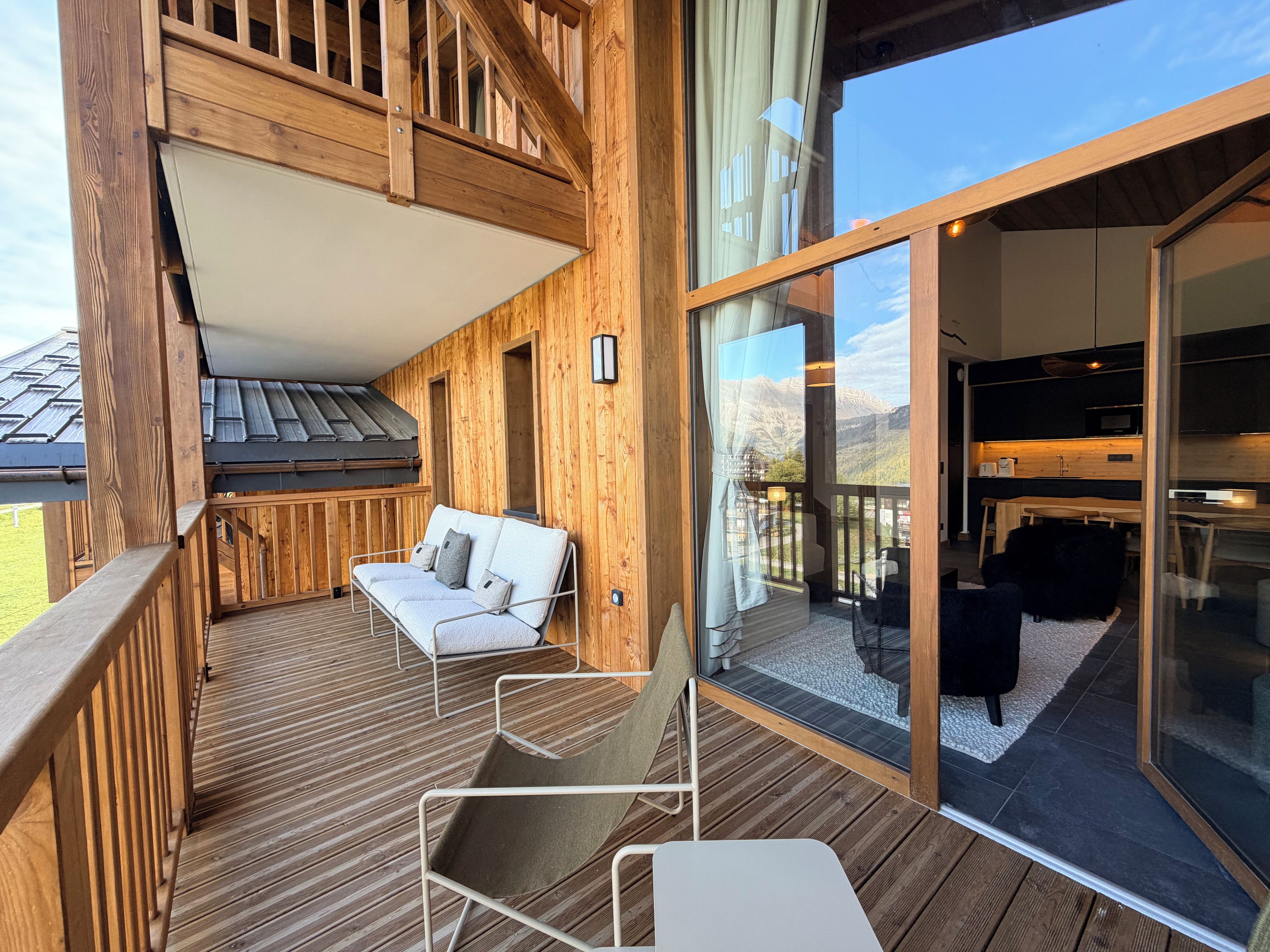A wooden deck with a sofa and chairs, a glass door leading to a kitchen, and a view of mountains.