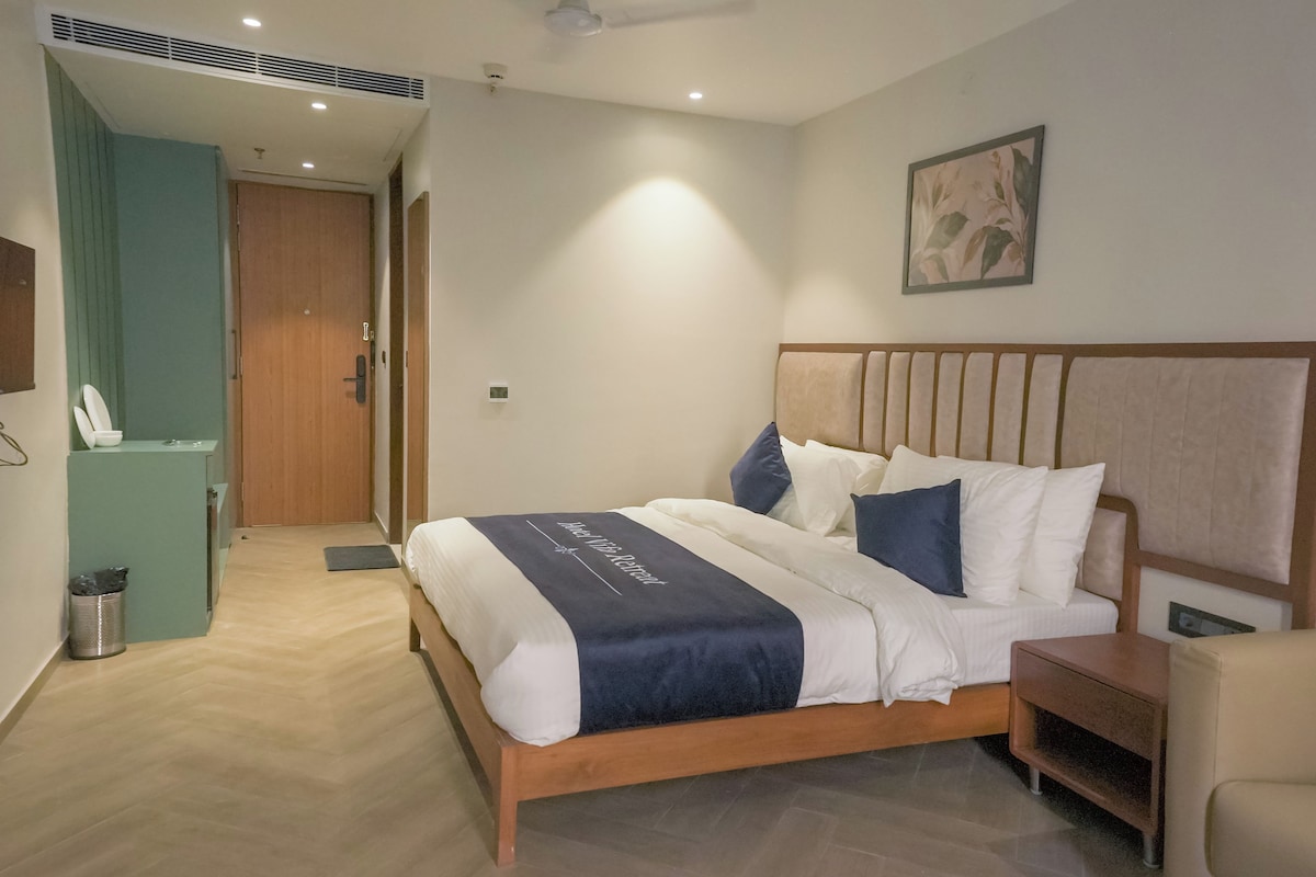 A hotel room with a wooden bed, a nightstand, a desk, and a chair. There is a door leading to another room and a trash can.