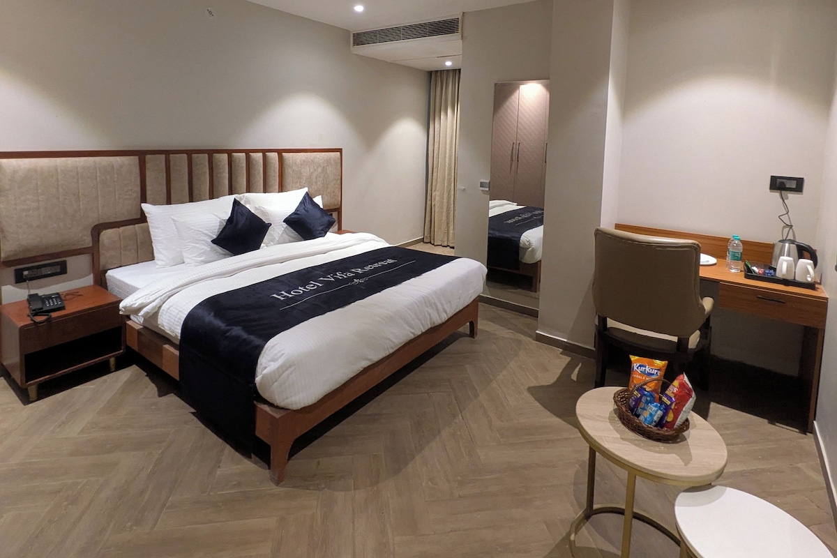 A hotel room with a bed, bedside table, desk, chair, and a small table with snacks.