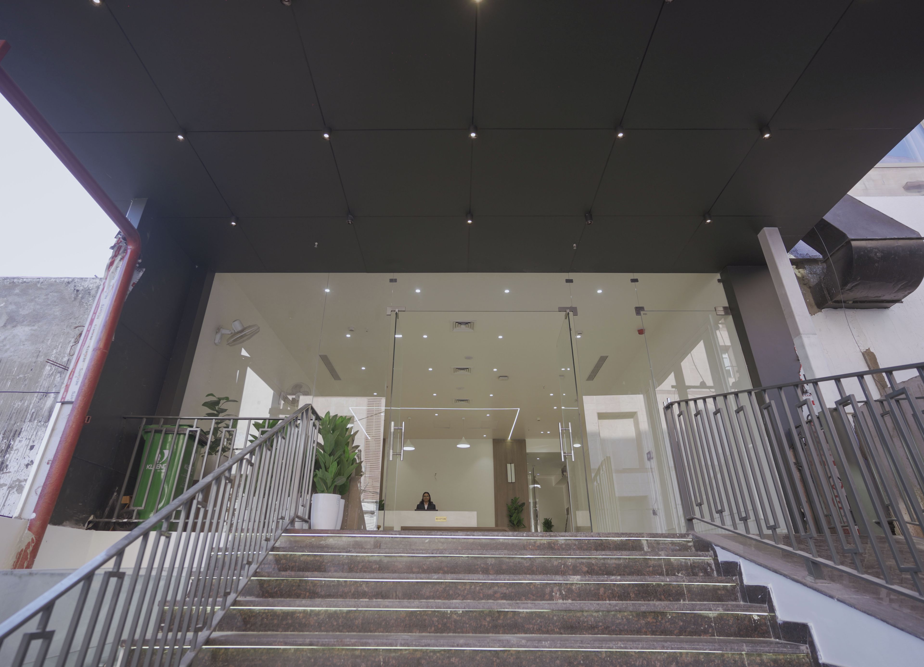 Una escalera conduce a un área de recepción con puertas de vidrio, barandilla metálica e iluminación empotrada en el techo.