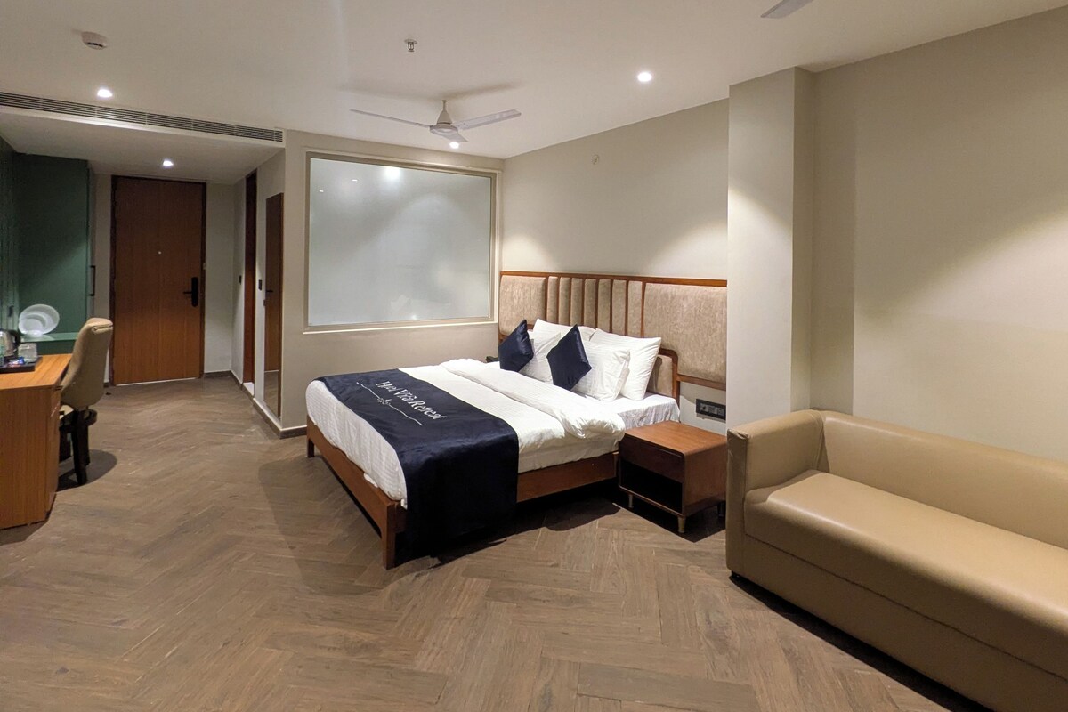 A hotel room with a bed, a desk, a chair, a sofa, a ceiling fan, and a large window.