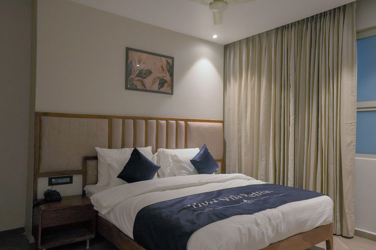 A hotel room with a bed, a nightstand, a painting, and a window with curtains.