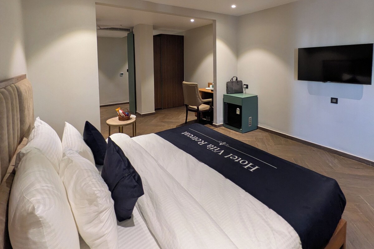 A modern hotel room with a large bed, a desk, a chair, a television, and a wardrobe.