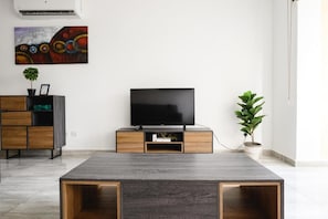 Ein modernes Wohnzimmer mit einem Flachbildfernseher auf einem Holzständer, einem Couchtisch in der Mitte und einer Topfpflanze rechts.
