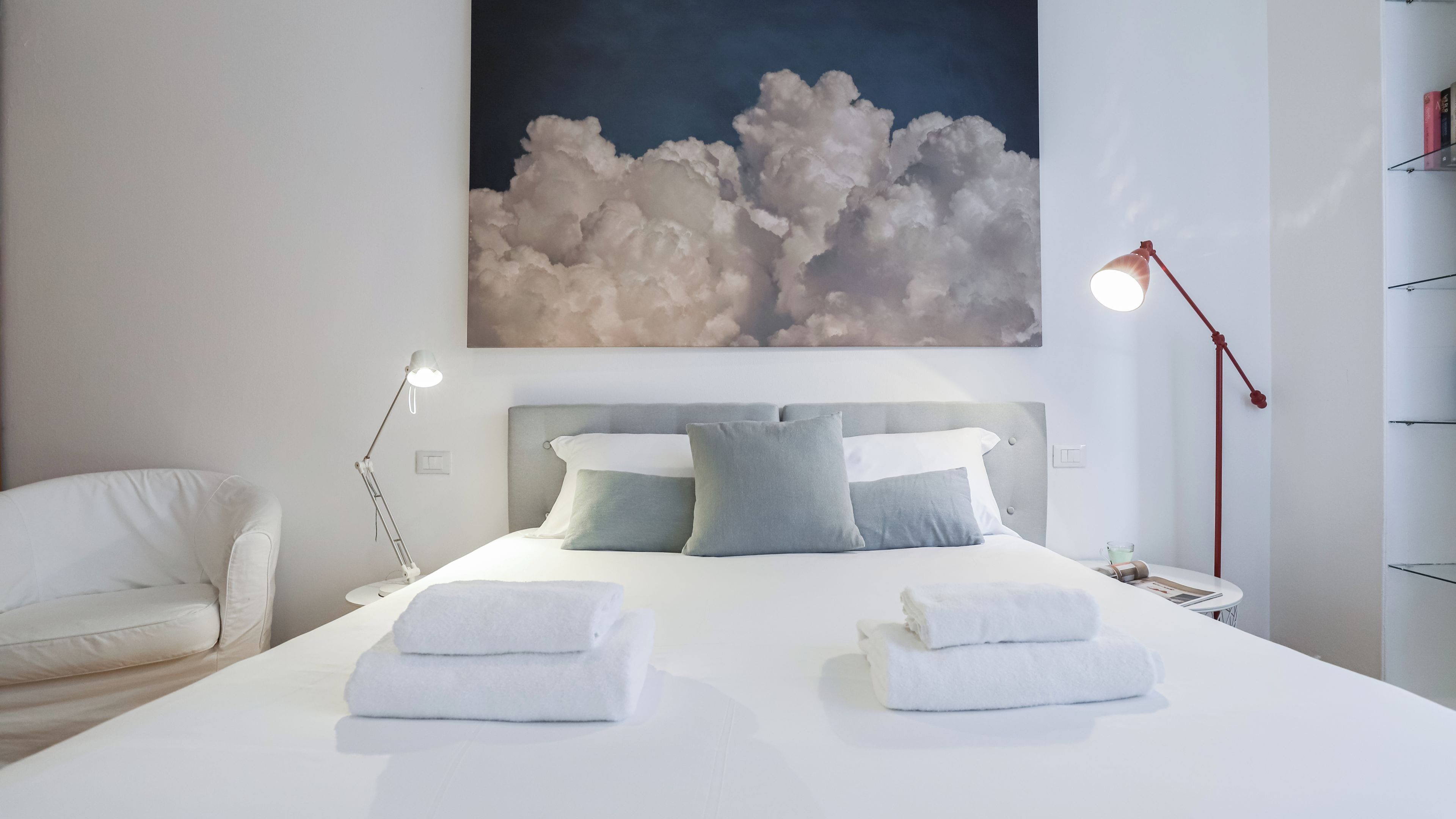 A modern bedroom with a large bed, two bedside lamps, a chair, and a painting of clouds on the wall.