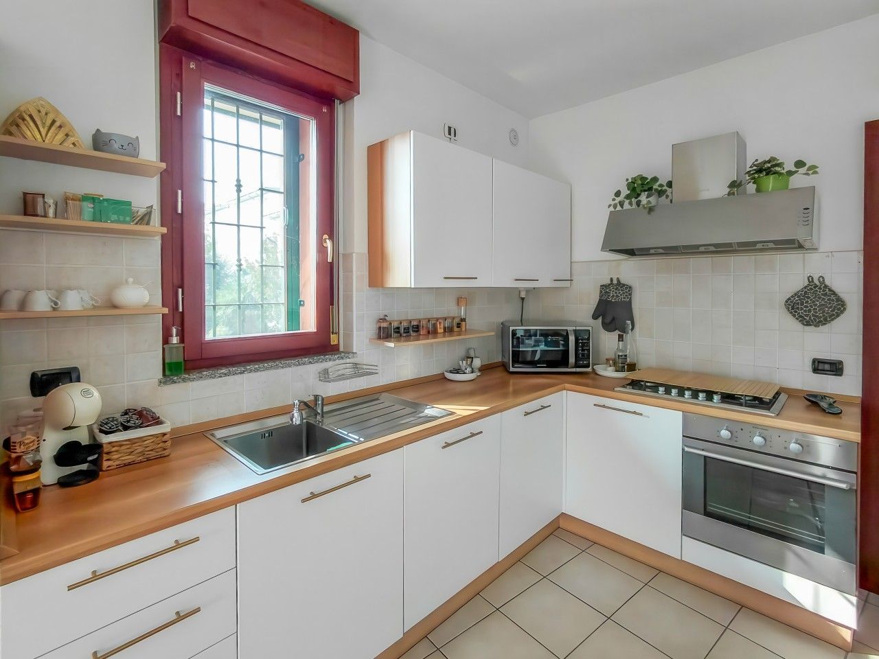 A modern kitchen with white cabinets, a stainless steel oven, a microwave, and a window with a red frame.