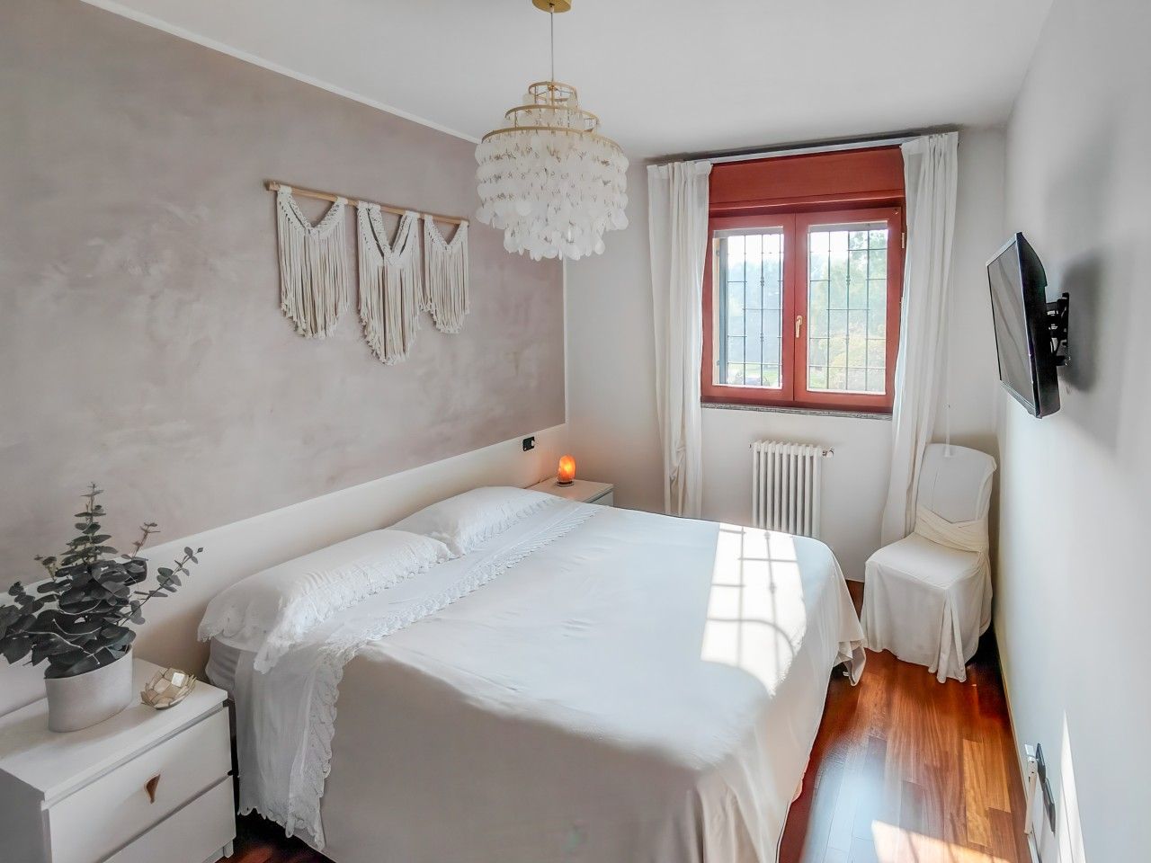 Ein Schlafzimmer mit einem großen Bett, Nachttischen, einem Schreibtisch, einem an der Wand montierten Fernseher, einem Fenster mit Vorhängen und einer Heizung.