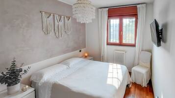 Ein Schlafzimmer mit einem großen Bett, Nachttischen, einem Schreibtisch, einem an der Wand montierten Fernseher, einem Fenster mit Vorhängen und einer Heizung.