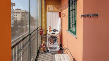 A narrow balcony with a washing machine, a drying rack, and a small window with a green frame.