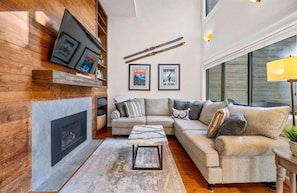 A living room with a fireplace, a sofa, a coffee table, and a TV mounted on the wall. - Spacious Sugarbush Mtn Retreat, Fireplace, Loft & Ski Room, Sleeps up to 8 (Warren)