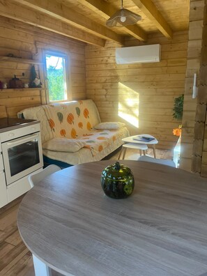 A cozy cabin interior with a wooden ceiling, a sofa with a pumpkin pattern, a small table, a decorative pumpkin vase, and a kitchenette with an oven. - New Listing (Céreste-en-Luberon)