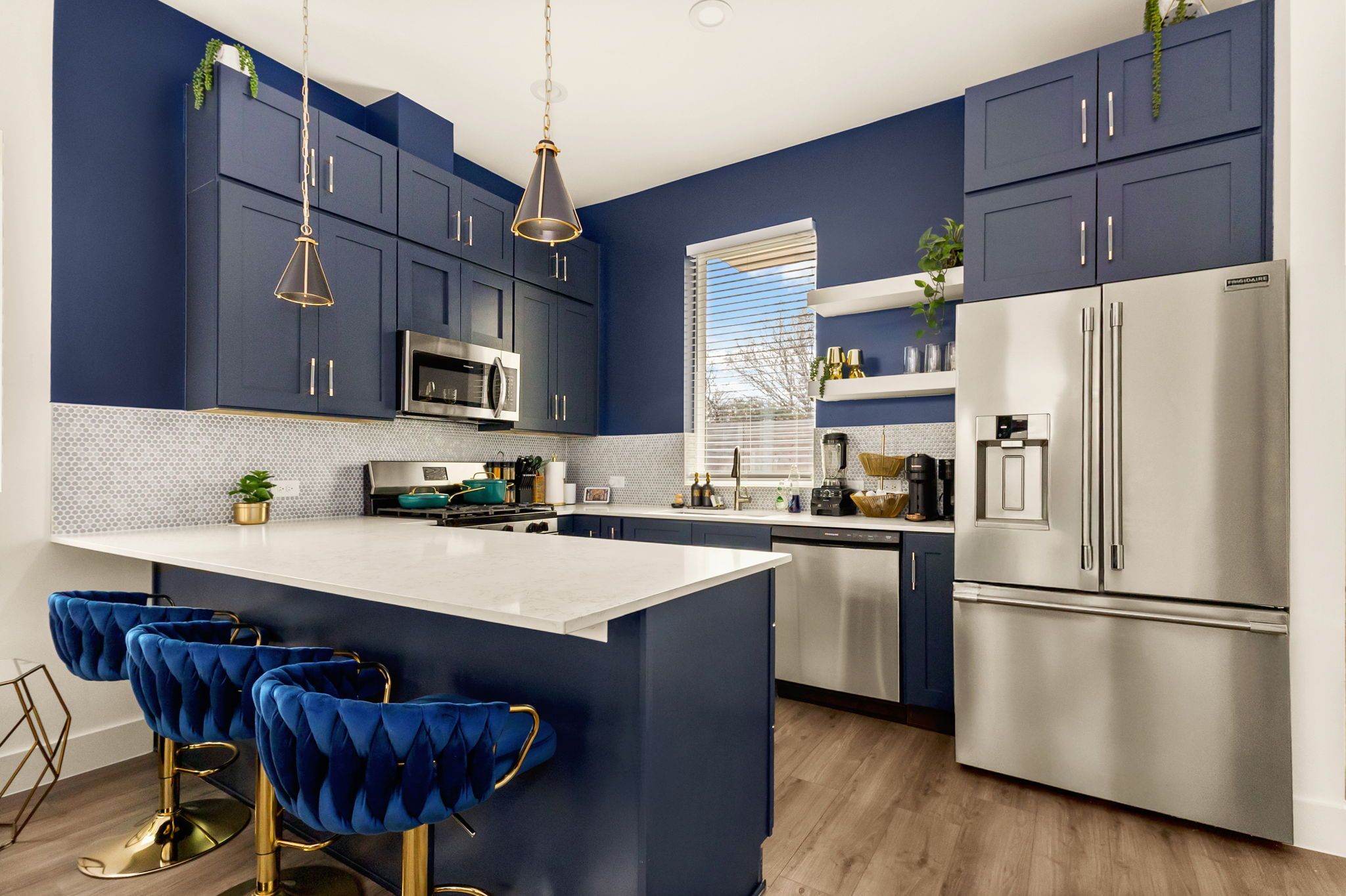 A modern kitchen with dark blue cabinets, stainless steel appliances, a white island with bar stools, and a window with blinds.