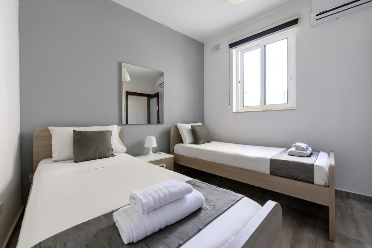 Et hotelværelse med to senge, et spejl, et vindue og et sengebord med en lampe.