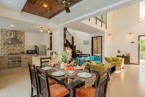 A spacious kitchen with modern appliances, a dining area with a wooden table and chairs, and a living room with a sofa and a staircase. - Staymaster Aqua Sol - 3BR Villas w Pool (Assagao)